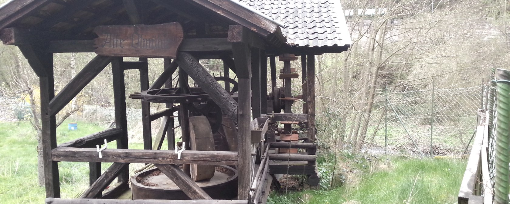 Eine alte hölzerne Ölmühle mit einem kleinen Dach steht in einer grünen, bewachsenen Umgebung., © Ti Hocheifel-Nürburgring
